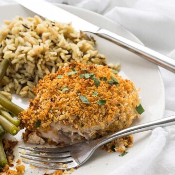 A cut piece of herb breaded chicken on a plate with a fork and knife.