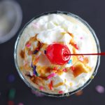 Overhead view of a shake in a glass topped with whipped topping, sprinkles and a cherry.