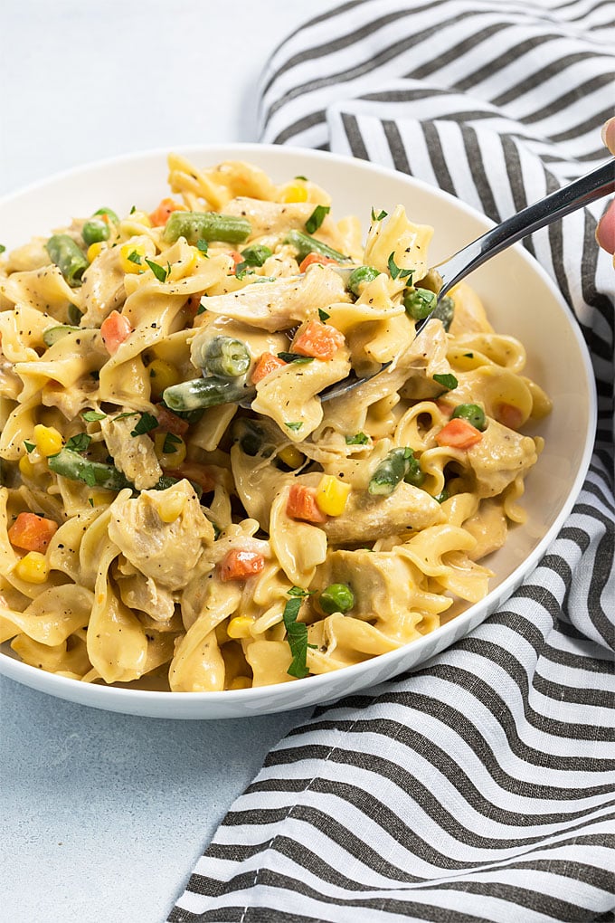 A spoon in a white bowl of cheesy noodles with chicken and vegetables.