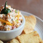 Pimento cheese topped with a jalapeno stem in a bowl surrounded by chips.