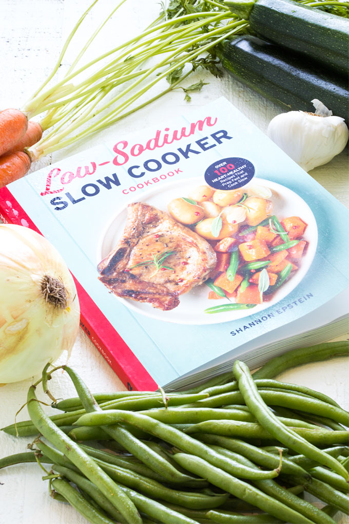 A cookbook on a white surface surrounded by fresh vegetables.