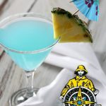 A blue cocktail in a martini glass garnished with pineapple and an umbrella.
