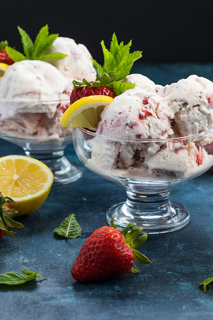 Front view of two glass bowls of ice cream garnished with strawberries, lemon and mint.