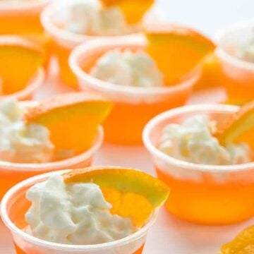 Orange jello shots topped with whipped cream and orange slices in small plastic cups.