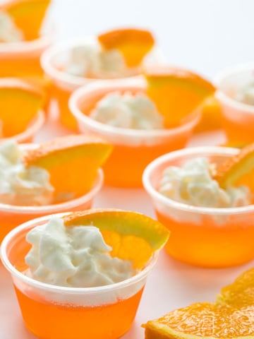 Orange jello shots topped with whipped cream and orange slices in small plastic cups.