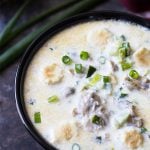Oyster stew topped with sliced green onions and soup crackers in a black bowl.