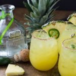 Three margaritas garnished with pineapple and jalapeno beside a bottle of tequila.