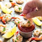 A hand dipping a shrimp into a stainless condiment cup of cocktail sauce.