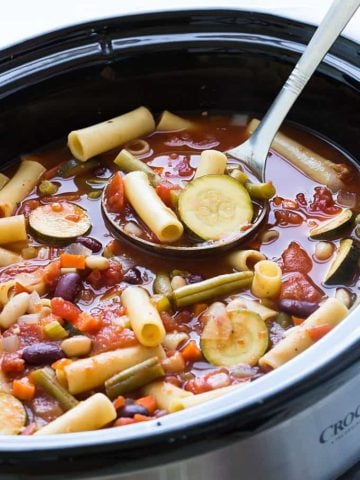 A stainless ladle ladling minestrone from an oval slow cooker.