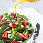 Pouring vinaigrette on a salad with strawberries and spinach in a white bowl.