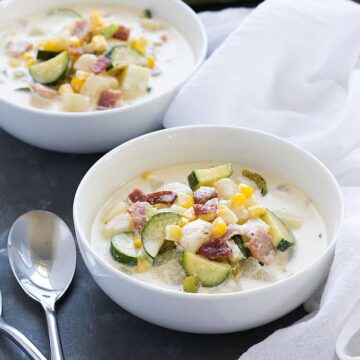 Two white bowls of zucchini and corn chowder beside two spoons.