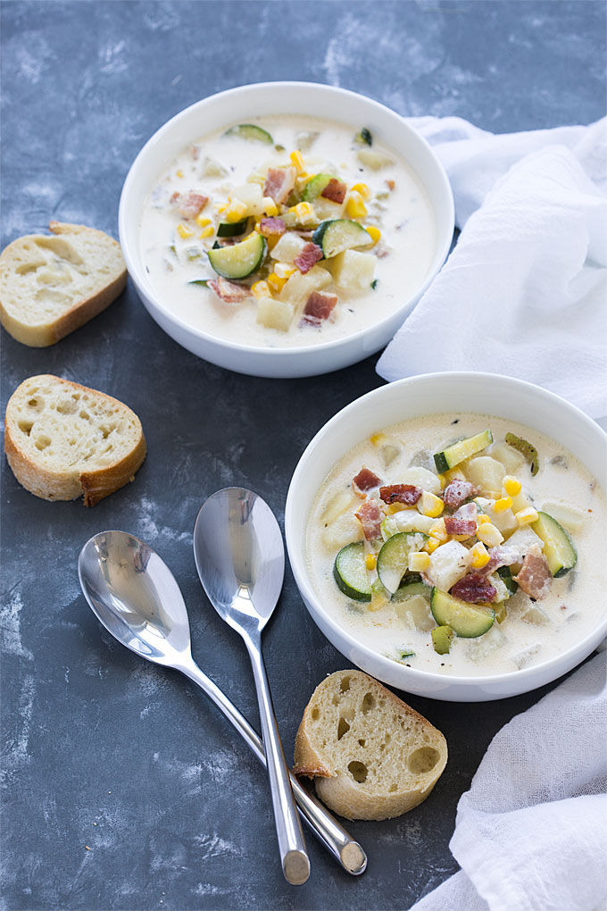 Overhead view of two white bowls of chowder by sliced French bread and two spoons.