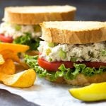 Two fish salad sandwiches by potato chips and a pickle spear on parchment paper.
