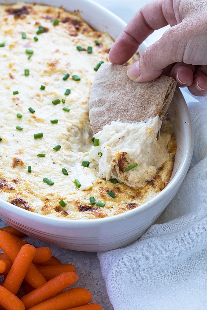 A hand dipping a piece of pita bread into a dish of crab dip.