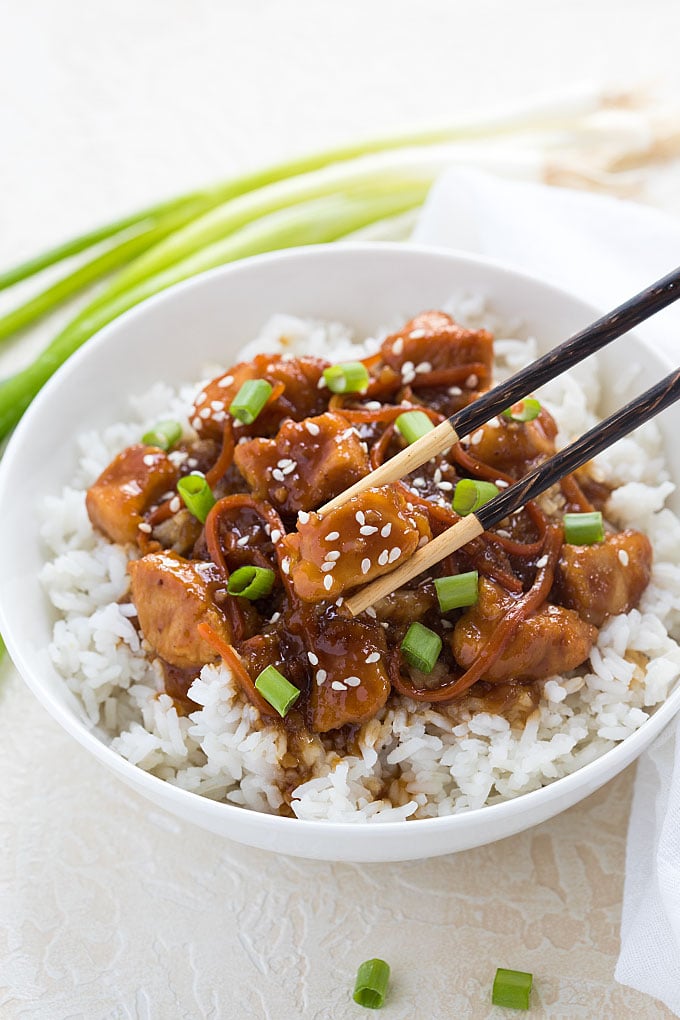 Mongolian chicken topped with green onions over rice in a  bowl with a pair of chopsticks.