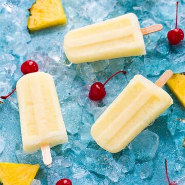 Overhead view of three yellow popsicles on a blue surface with ice, cherries and pineapple.