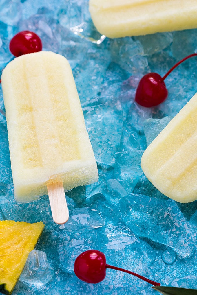 A closeup of a popsicle on a blue surface with ice, cherries and a pineapple slice.