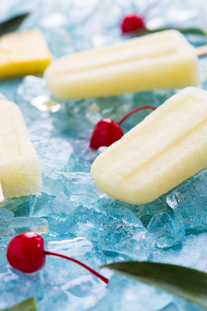 Closeup view of a pina colada popsicle on a blue surface with ice and cherries.