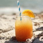 Front view of an orange cocktail with a straw in the sand on a beach.