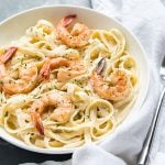 A white bowl of pasta with shrimp and dill by a white napkin.