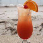 An orange cocktail in a hurricane glass sitting in the sand on a beach.