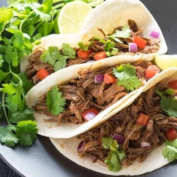 A closeup of three soft tacos with Mexican shredded beef on a black plate with cilantro.
