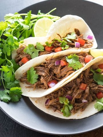 A closeup of three soft tacos with Mexican shredded beef on a black plate with cilantro.