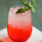 A pink cocktail in a glass with ice garnished with fresh mint.