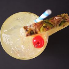 Overhead view of a yellow cocktail garnished with pineapple and a cherry.