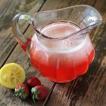 Strawberry lemonade in a glass pitcher on a wooden surface.