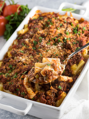 A serving spoon removing baked ziti with ground beef from a white baking dish.