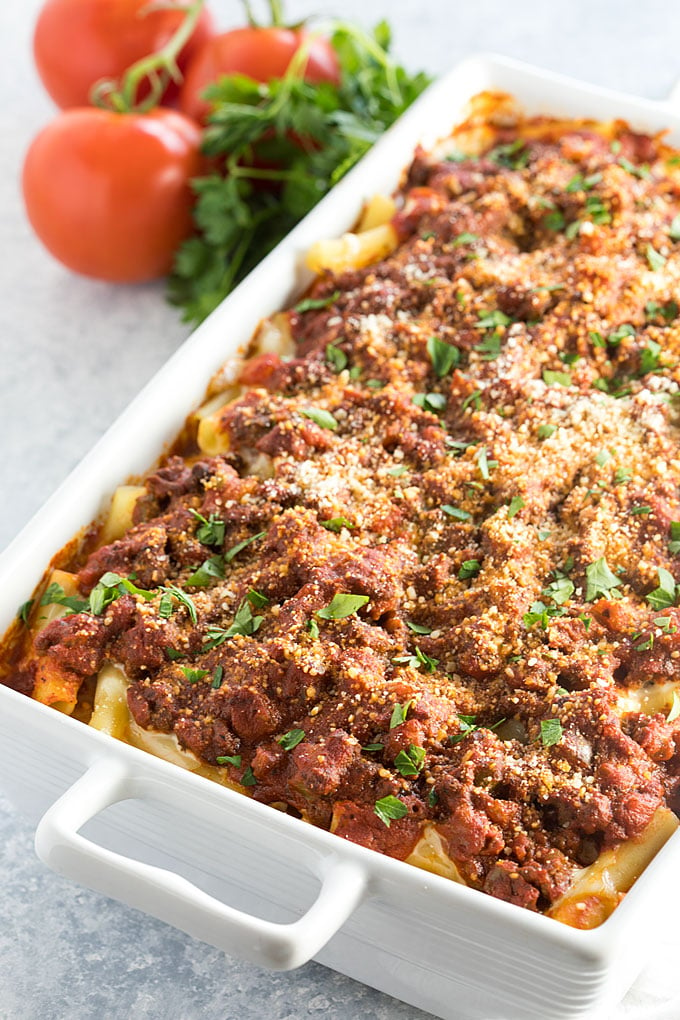 Baked ziti in a rectangle white baking dish beside parsley and tomatoes.