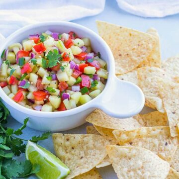 Salsa with cucumbers, tomatoes and onion in a white bowl beside tortilla chips.