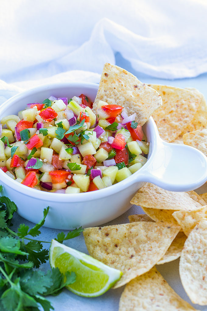 A tortilla chip in a white bowl of cucumber salsa topped with fresh chopped cilantro.