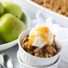Caramel syrup being poured over apple crisp topped with vanilla ice cream in a white ramekin.