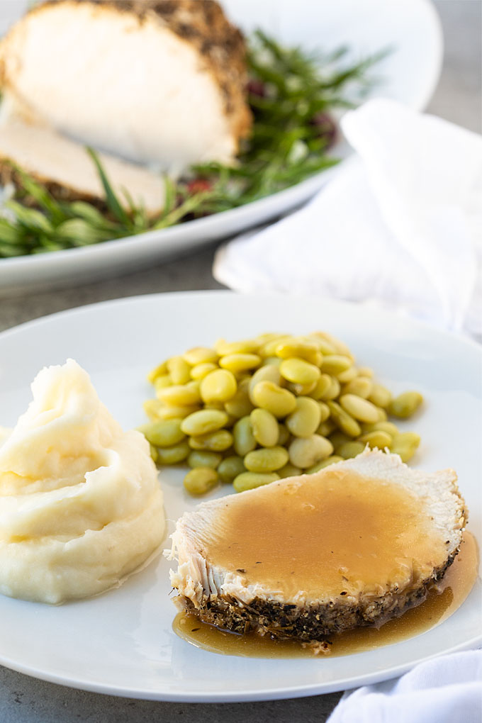A slice of turkey breast with gravy, lima beans and mashed potatoes on a white plate.