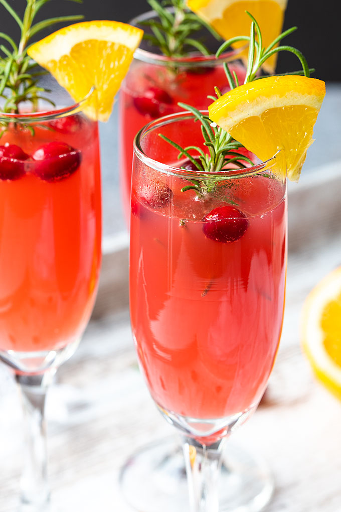 A closeup of a mimosa garnished with rosemary, fresh cranberries and an orange slice.