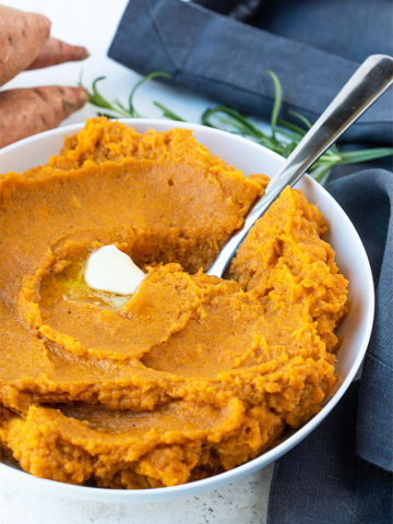 A white bowl of mashed sweet potatoes with a pat of butter and a spoon.