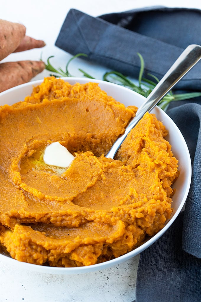 A white bowl of mashed sweet potatoes with a pat of butter and a spoon.