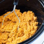 A potato masher mashing sweet potatoes in an oval slow cooker with overlay text.
