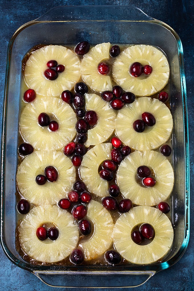 Sliced pineapple and cranberries over a brown sugar mixture in a baking dish.
