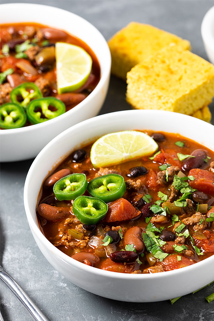 Two white bowls of chili beside slices of baked cornbread.