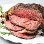 A sliced boneless prime rib on a white serving platter garnished with fresh rosemary and cranberries.