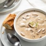 Cream of mushroom soup in a white bowl on a plate with a spoon and a slice of French bread