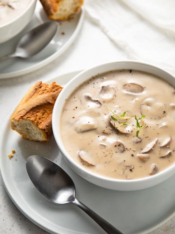 Cream of mushroom soup in a white bowl on a plate with a spoon and a slice of French bread