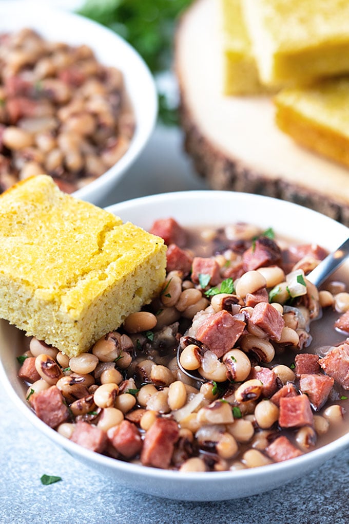 Black-eyed peas and ham in a white bowl with a spoon and a slice of cornbread.
