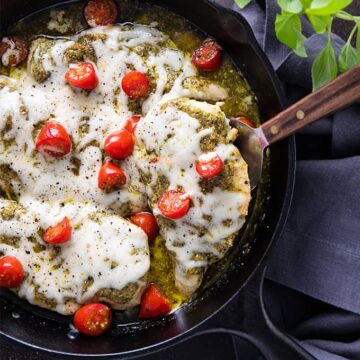 Baked pesto chicken topped with melted provolone cheese and cherry tomatoes in a cast iron skillet.