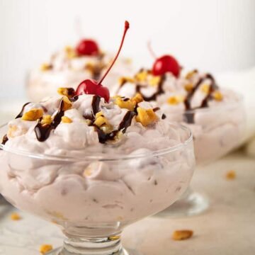Banana split fluff salad topped with chocolate syrup, walnuts and a cherry in a glass dessert dish.