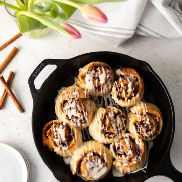 Glazed cinnamon rolls in a cast iron skillet beside a stack of small round white plates, cinnamon sticks, a vase of pink tulips and a gray striped kitchen towel.