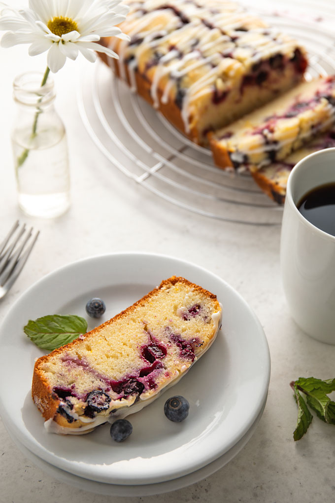 A slice of lemon blueberry bread on a round white plate with a sprig of mint, a white cup of coffee and 3 forks.